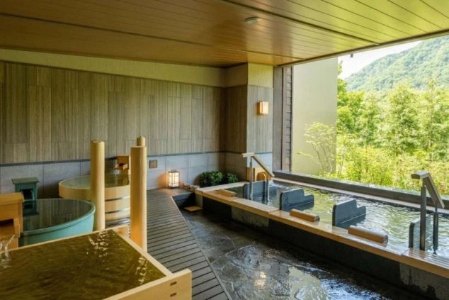 定山渓温泉の浴場。新緑の山々を望む大きな窓と、木製の浴槽が並ぶ開放的な露天風呂。