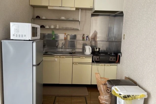  冷蔵庫や電子レンジが置かれた共用キッチン。シンク、ガスコンロ、棚があり、ゴミ箱とゴミ袋が床に置かれている。