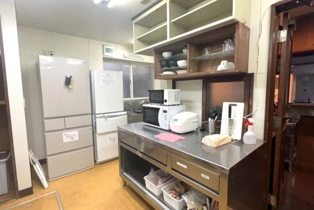 共用のキッチンコーナー。ステンレス製の調理台の上に、炊飯器、電子レンジ、トースターが並び、奥には大小2台の冷蔵庫が設置されている。
