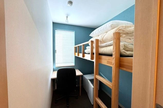 青い壁の部屋に、木製の二段ベッドが置かれ、ベッドの上には布団が積まれている画像。手前には勉強机と椅子、小さな冷蔵庫が見え、リゾートバイトの個室寮で、プライベート空間が確保された寝室のイメージ。