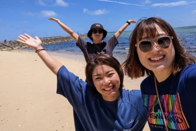 晴れた日のビーチで、笑顔で両手を広げる男女3人のセルフィー。