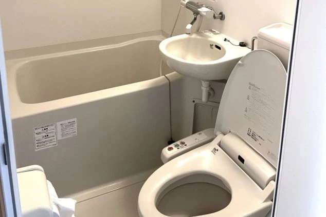 寮のユニットバス。手前に温水洗浄便座付きトイレ、奥に洗面台と浴槽が見える。壁に注意書きのステッカーが貼られている。