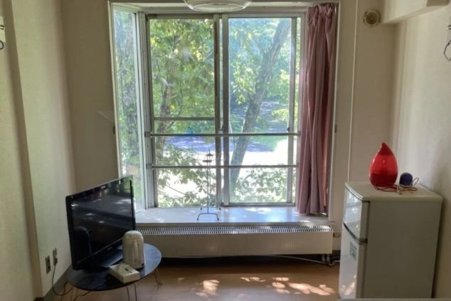 リゾートバイトの寮の部屋の画像。大きな窓から緑豊かな景色が見え、テレビと冷蔵庫が置かれている。
