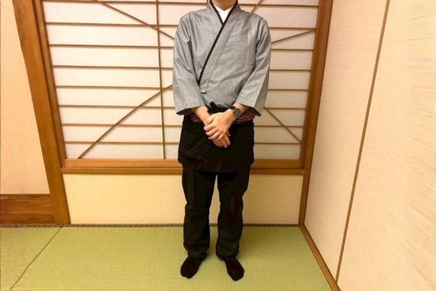 ハンガーに掛けられた、吉祥のレストランスタッフの制服。朱色（またはオレンジ色）の和風上着と、裾に波柄がデザインされた紺色の巻きスカートまたはエプロン。