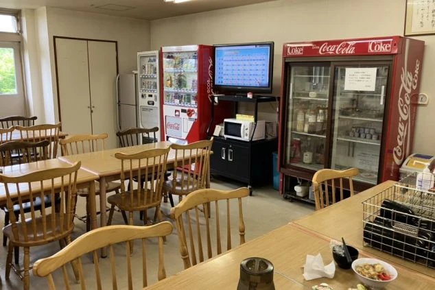 テーブルと椅子が並んだ社員食堂。部屋の奥には、自動販売機、冷蔵庫、電子レンジ、テレビが置かれている。