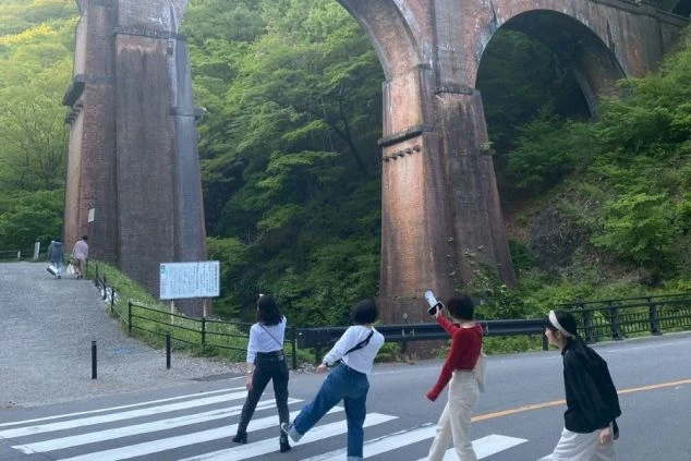 群馬・碓氷第三橋梁（めがね橋）を背景に、横断歩道を渡りながら笑顔で撮影を楽しむ4人の女性グループ。軽井沢エリアのリゾートバイトで出会った仲間との休日風景。