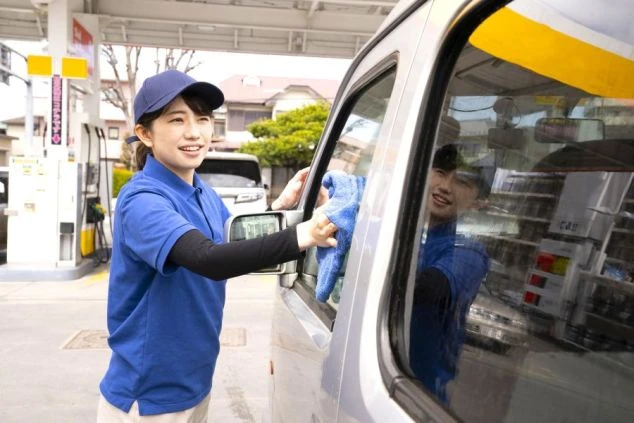 笑顔で車の窓を拭くガソリンスタンドの女性スタッフ。リゾート地のサービスステーションやレンタカー受付での住み込みバイト募集。