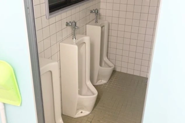 寮の共用トイレに設置された3台の壁掛け式小便器。床と壁はタイル貼りで、窓から光が差し込む明るい空間。清掃用具も置かれ、手入れされている様子。