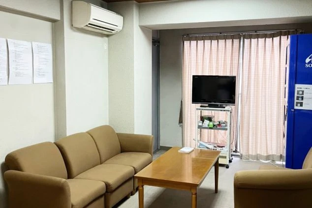 共用のリラックススペース。落ち着いた茶色のソファと木製テーブルが置かれ、奥にはテレビと自動販売機が設置されている。窓にはピンク色のカーテンがかかっている。