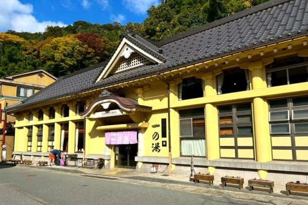 城崎温泉の七外湯の一つ「一の湯」の外観。江戸時代の歌舞伎座を彷彿とさせる桃山様式の黄色い建物と、紫色ののれんが特徴的な、リゾートバイト中の休日観光や湯巡りに人気のスポットを写した画像。