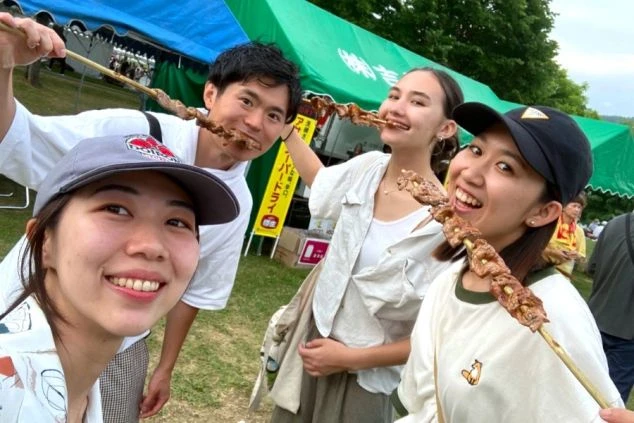 北海道のリゾートバイトで出会った仲間たちと、休日に地元のイベントを楽しむスタッフの画像。大きな串焼きを頬張る笑顔の男女4人組。