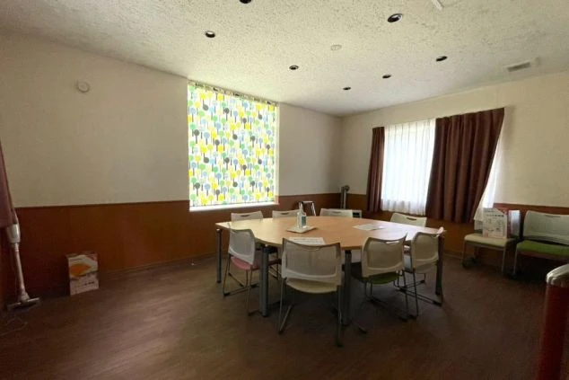 ワクトリの社員が出張で撮影した、共用リビングの画像。大きなテーブルと椅子が複数配置され、窓にはカラフルなブラインドとカーテンがかかっている。掃除機や消毒液も見える。