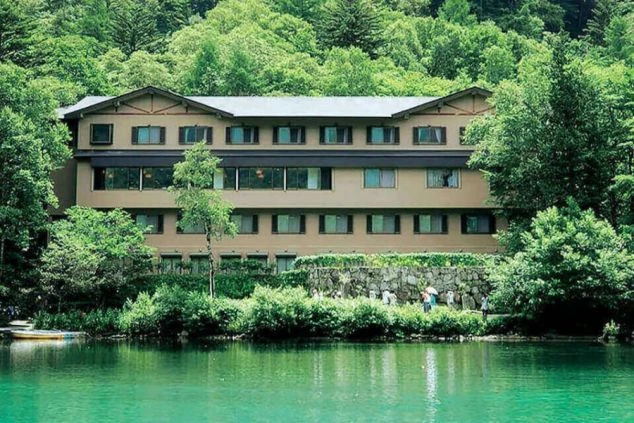 穏やかな水面に映し出される、緑豊かな森の中に佇む静かなホテルの外観。周囲の深い緑と調和した落ち着いた建物が、上高地の自然豊かな環境を感じさせる風景。