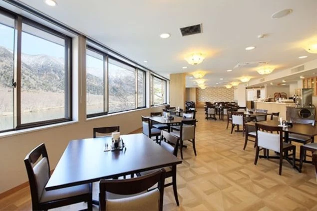 大きな窓から残雪の残る山々と梓川の絶景を望む、明るく広々としたホテルのレストラン内。木目調のテーブルが並び、景色を楽しみながら食事ができる開放的な空間。