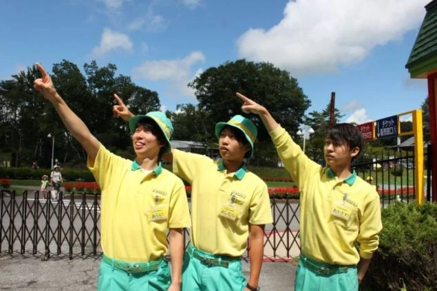 黄色いポロシャツに水色のズボンと帽子を着用した、リゾートバイトの制服姿の男性3人の画像。屋外で空を指さしています。晴れた空と緑の木々が見え、テーマパークやレジャー施設での仕事風景を表しています。
