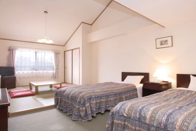 和洋室の客室。手前には2台のシングルベッドが並び、奥の畳スペースにはローテーブルと座布団、テレビが配置されている。天井は高く開放感がある。