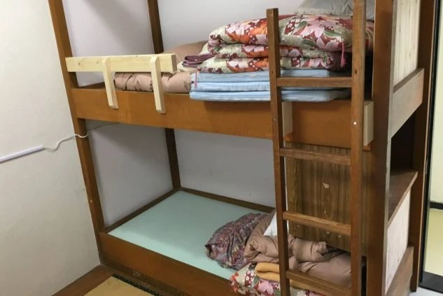 木製の2段ベッド。上段には花柄の布団が積まれ、下段には水色のマットレスと枕、掛け布団が置かれている。枕元には木製のはしごが掛かっている。