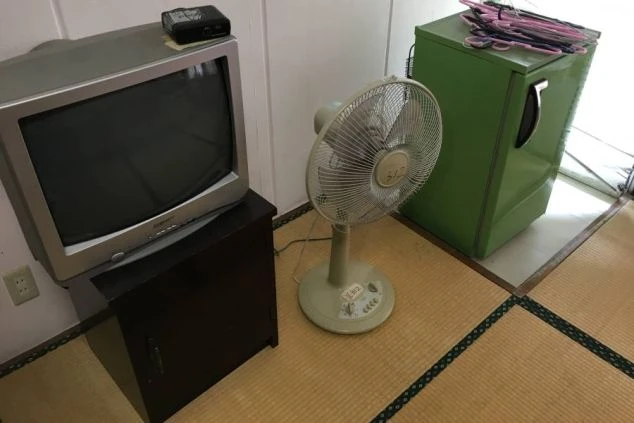 畳の部屋の隅。黒い台の上に置かれた古いブラウン管テレビ、ベージュの扇風機、緑色の小型冷蔵庫が並んでいる。冷蔵庫の上にはハンガーが置かれている。