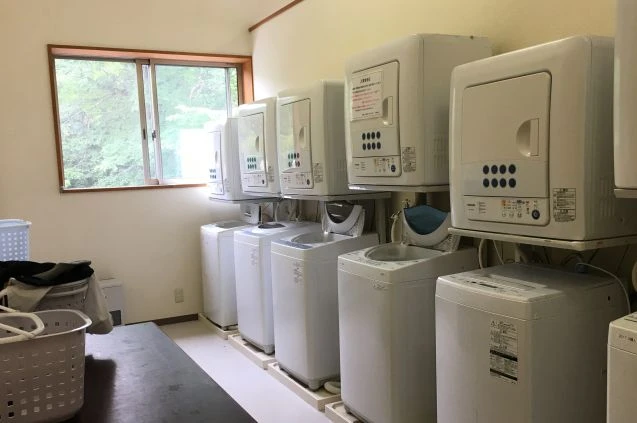 共用の洗濯室。白い縦型洗濯機の上に乾燥機が設置されたユニットが、壁に沿って5台ほど並んでいる。手前には洗濯物が入ったカゴとテーブルがある。