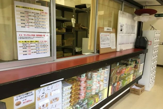 売店のカウンター。ショーケースにはカップ麺や菓子が並び、ガラス窓にはパンの販売メニューが貼られている。奥の棚には段ボール箱が保管されている。