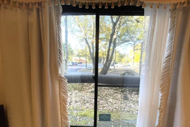 客室の窓からの眺め。白いレースのカーテン越しに、駐車場と秋の色づき始めた木々が見える。