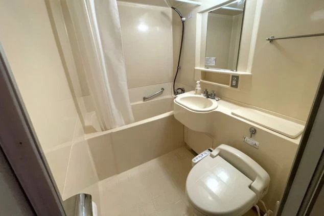 清潔感のあるコンパクトなユニットバス。白いシャワーカーテンが引かれ、洗面台の鏡が正面に見える。