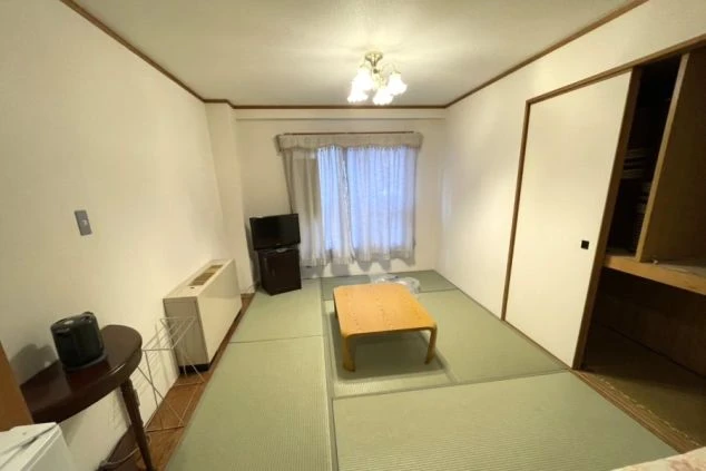 畳敷きの和室。中央にローテーブルがあり、奥の窓には白いカーテンがかかっている。