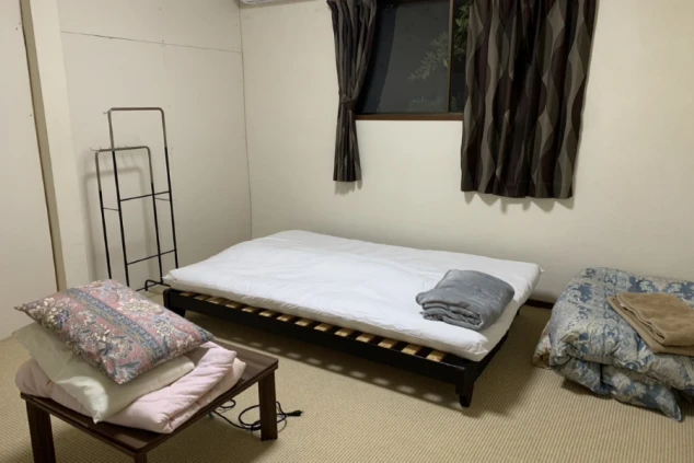 ワクトリの社員が出張で撮影した、リゾートバイトの寮の部屋の画像。畳敷きの部屋にシンプルなベッドと布団が置かれ、窓には遮光カーテンがかかっている。ハンガーラックもある。