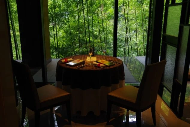 竹林を望む幻想的な高級リゾートホテルのレストラン席。落ち着いた照明の中、窓の外に広がる緑豊かな竹林を眺めながら食事が楽しめる、贅沢な職場環境のイメージ。