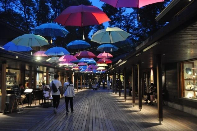 夜の帳が下りた長野県軽井沢町の「ハルニレテラス」で、ライトアップされたカラフルなアンブレラスカイの画像。ウッドデッキの街並みを歩く人々を優しく照らす傘の灯り。