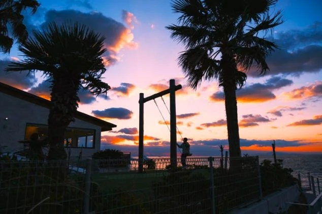 路島の海岸沿いにある「幸せの階段」付近の大きなブランコ。美しい夕焼け空とパームツリーを背景に、リゾート感あふれる黄昏時の風景。