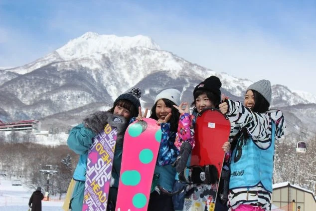雄大な雪山を背景にボードを持ってポーズをとるスタッフ。冬の思い出作りと貯金が両立できるスキー場リゾートバイト。