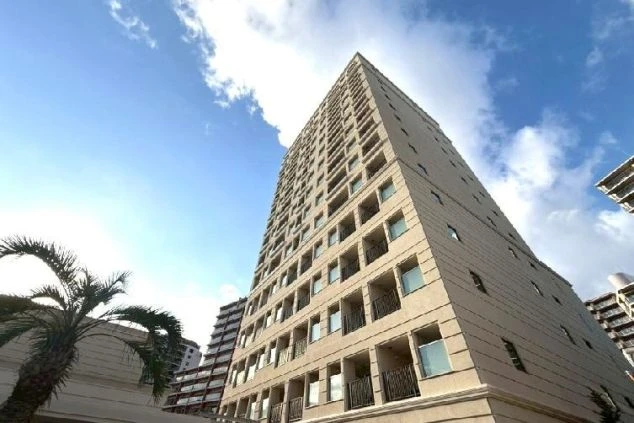 青空に向かってそびえ立つ、モダンでスタイリッシュな高層ホテルの外観。淡いベージュの外壁に規則正しく並ぶベランダが特徴的で、手前には南国情緒を感じさせるヤシの木が配された、開放感のある風景。