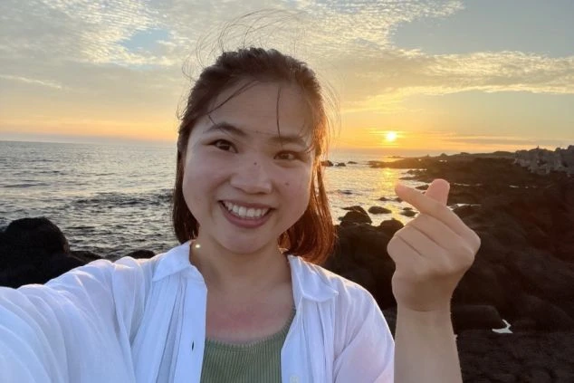 水平線に沈む美しい夕日をバックに笑顔で指ハートを作るリゾートバイトの女性スタッフの画像。