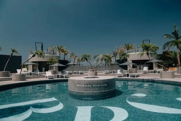 ヤシの木とパラソルに囲まれた「BOTANICAL POOL CLUB」の屋外プール。中央に円形の焚き火台のようなオブジェがあり、奥にはバーカウンターを備えたモダンで贅沢なリゾート空間。