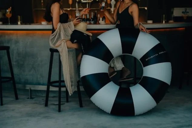 夜のプールサイドバーで寛ぐ水着姿の女性たち。手前には大きな白黒ストライプの浮き輪が置かれ、カウンターでドリンクを手に楽しむ、大人のリゾートの夜を象徴するスタイリッシュな一枚。