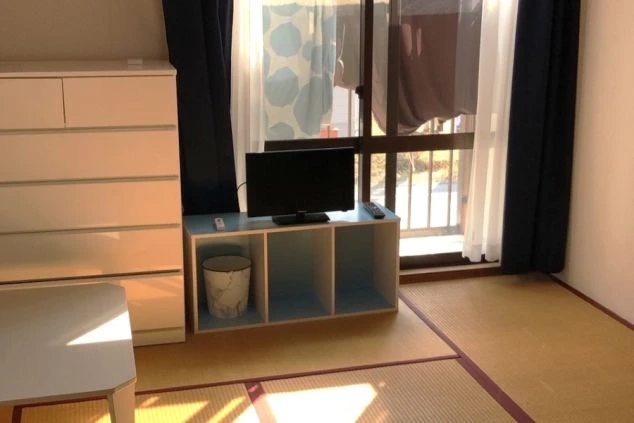 畳敷きの落ち着いた和室の寮自室。窓際の中座卓には小型テレビが置かれ、横には白い多段チェストを配置。掃き出し窓からは外光が差し込み、ベランダに干された洗濯物が透けて見える生活感のある空間。