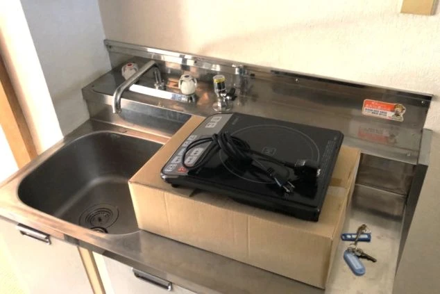 寮の自室に備え付けられたシンプルなミニキッチン。ステンレス製のシンク横には、段ボールの上に置かれた卓上IHクッキングヒーターがあり、自炊が可能な設備。手前には部屋の鍵も置かれている。