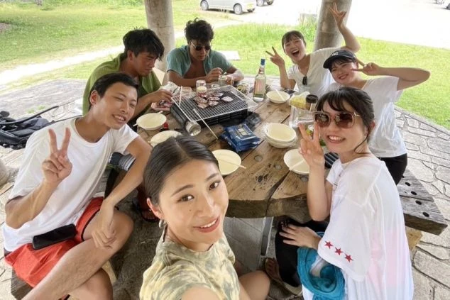 緑に囲まれた屋外の円卓で、コンロを囲みお肉を焼きながらBBQを楽しむ男女グループ。満面の笑みでピースをして自撮りをする、ワクトリが紹介する充実したリゾートバイトの休日風景です。