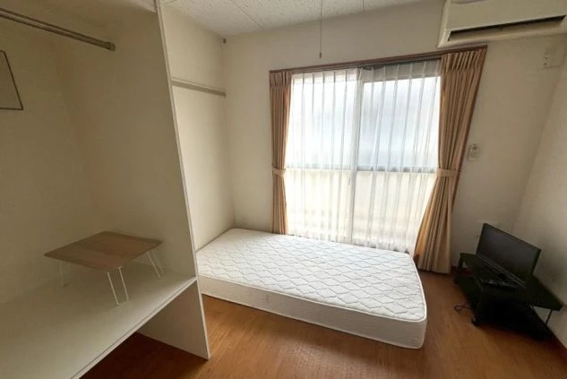 フローリングの床にマットレスが直置きされたシンプルな寮の個室。大きな掃き出し窓からは自然光が入り、壁際の棚や小型テレビ、エアコンなど生活に必要な最小限の設備が整っている。