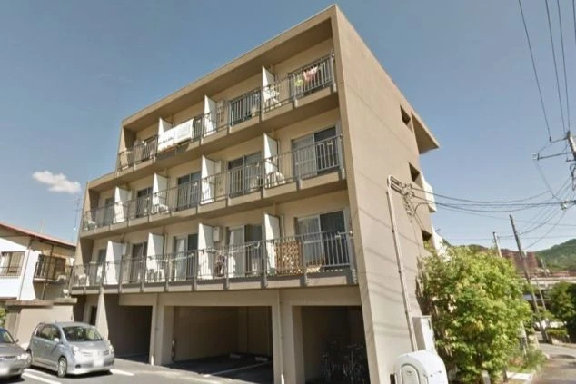 ベージュの外壁に各部屋のベランダが並ぶ、4階建ての寮の外観。1階部分はピロティ形式の駐車場になっており、建物の横には植栽が整えられた、日当たりの良い落ち着いた佇まい。