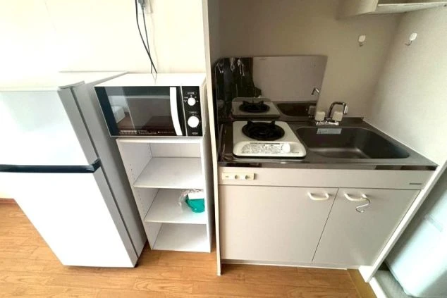 居室内に備え付けられたコンパクトなキッチン。1口ガスコンロとシンクの横に、小型冷蔵庫、電子レンジ、収納棚が効率よく配置されており、限られたスペースで自炊が可能な構成。