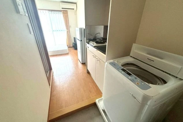 入り口から居室の奥までを見渡した寮の室内風景。手前には全自動洗濯機が置かれ、奥にはキッチン、さらにその先にはバルコニーへと続く居住スペースが広がる、機能的なワンルームのレイアウト。