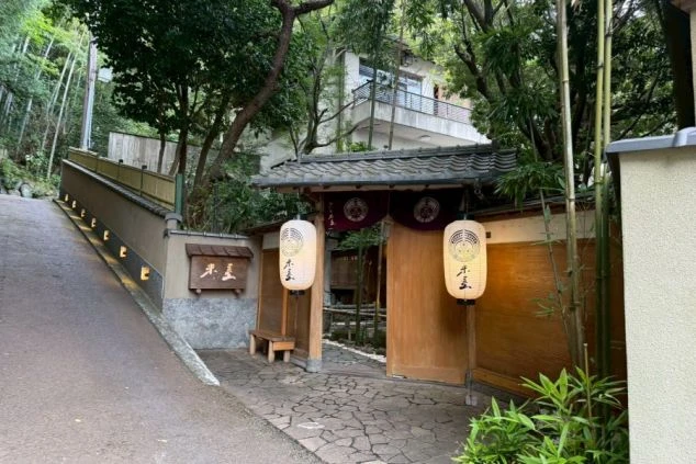 豊かな緑に囲まれた老舗旅館の重厚な門構え。店名の入った提灯が暖かく灯り、石畳の通路や竹林が静寂で格式高い和の風情を演出している。落ち着いた高級感の漂う玄関アプローチの風景。