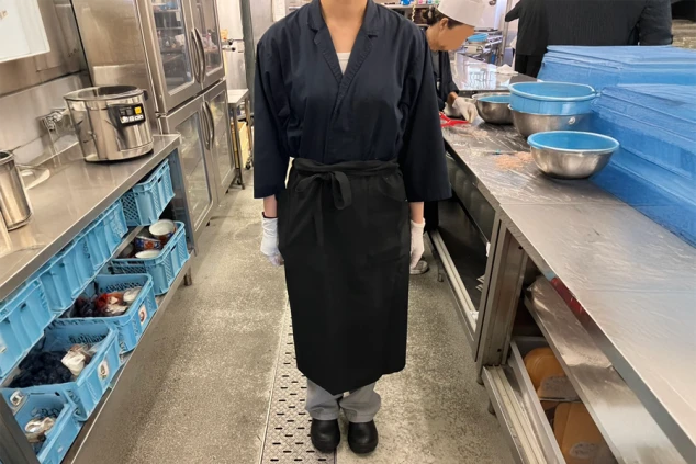 厨房で、黒い調理服とエプロンを着用した調理補助スタッフ。白い手袋をはめている。