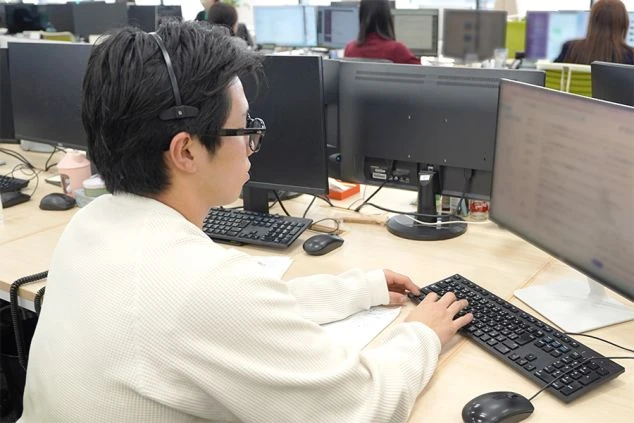 複数のモニターが並ぶオフィスで、ヘッドセットを装着し、真剣な表情でキーボード入力を行う男性社員。デスクワークに集中する、IT企業の日常的な業務風景を捉えた一枚。82文字