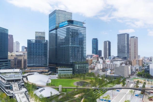 青空の下にそびえ立つ、複数階建ての大型ホテルの外観。手前には「平成館 しおさい亭 別館 花月」と書かれた大きな看板と、植栽に囲まれた広々としたエントランスアプローチが広がっている。